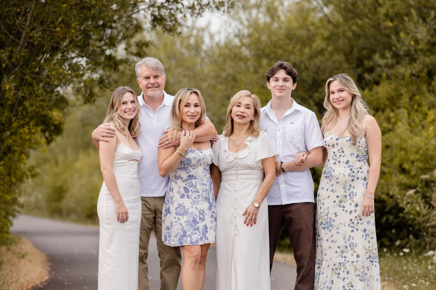 A group wearing coordinating white and floral outfits poses together on a tree-lined outdoor path.