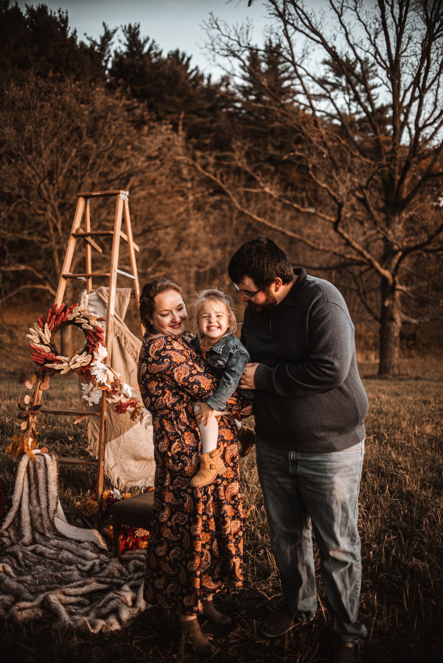Cozy autumn outdoor family photo with wooden ladder and blankets in a rustic woodland setting at sunset.