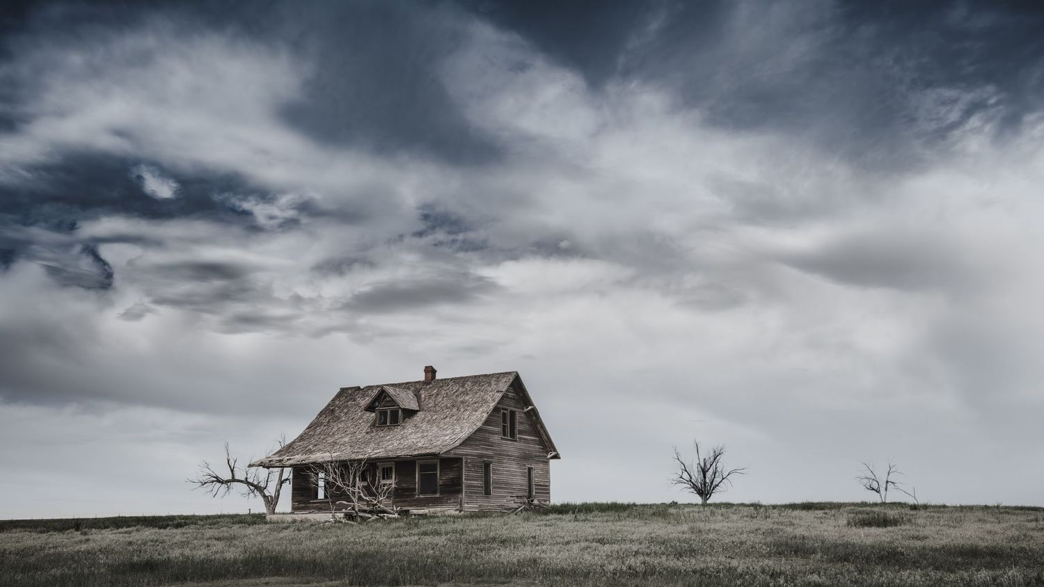 Abandoned wooden farmhouse stands alone on prairie under moody storm clouds.
