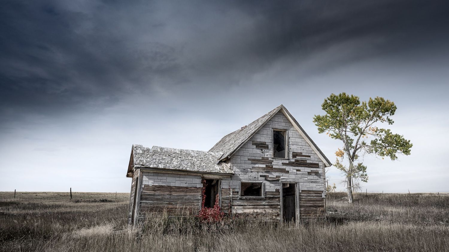 Weathered abandoned farmhouse with broken windows stands in open grassland under stormy sky.