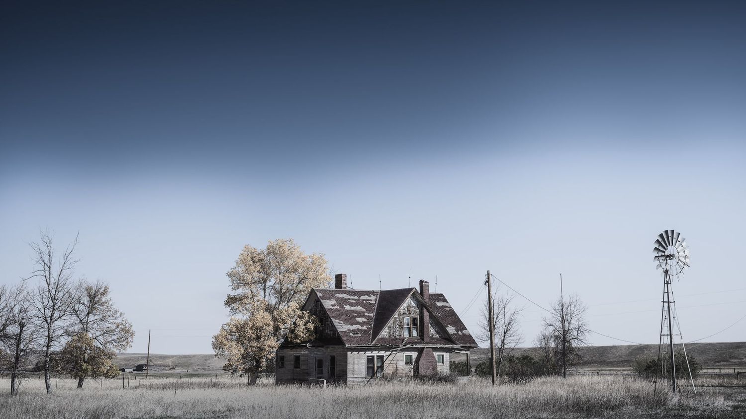 Abandoned farmhouse and windmill stand in winter field under muted sky.