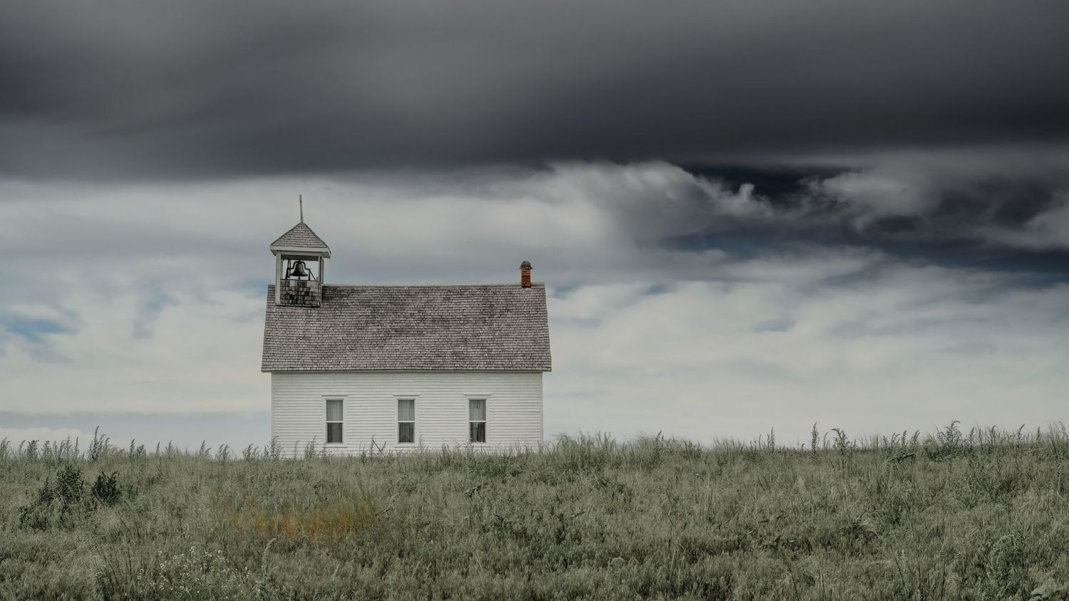 Small white prairie church with bell tower under threatening storm clouds.