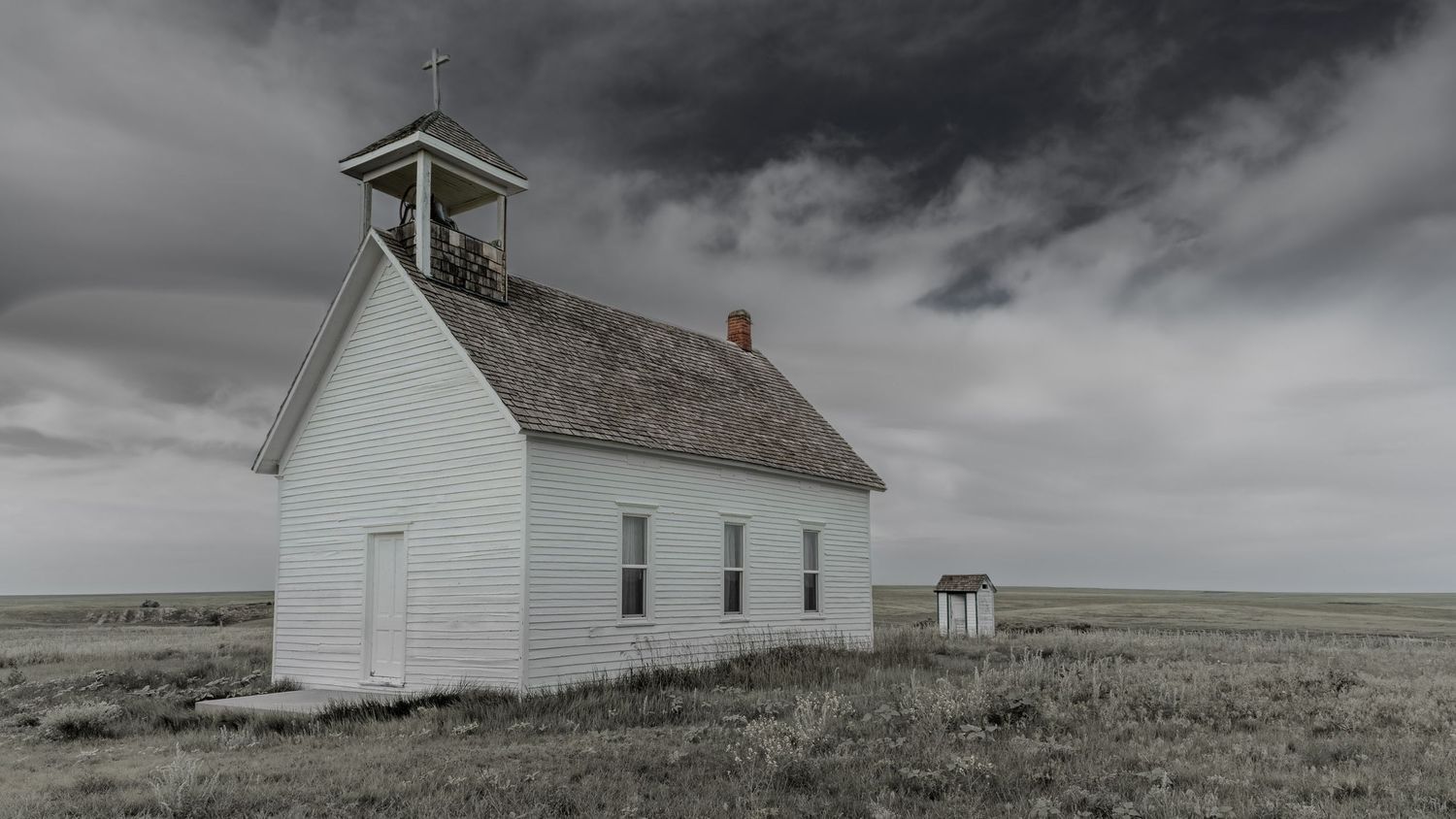 Historic white country church stands solitary on prairie under dark storm clouds.