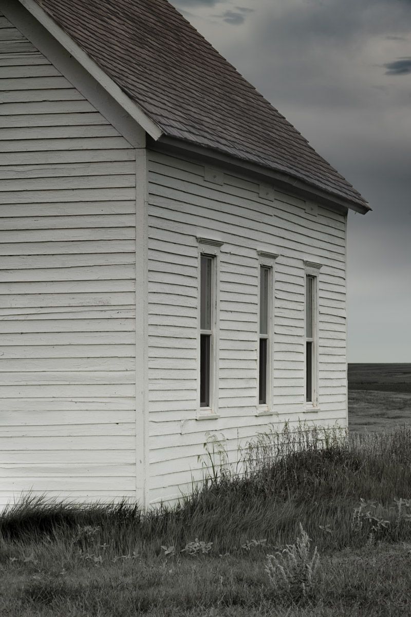 White clapboard building with three rectangular windows stands against a moody sky on the prairie.