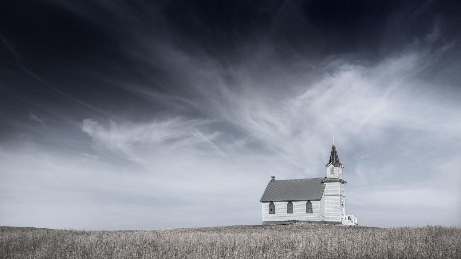 Small white prairie church with steeple sits alone on grassy hill under cloudy sky.