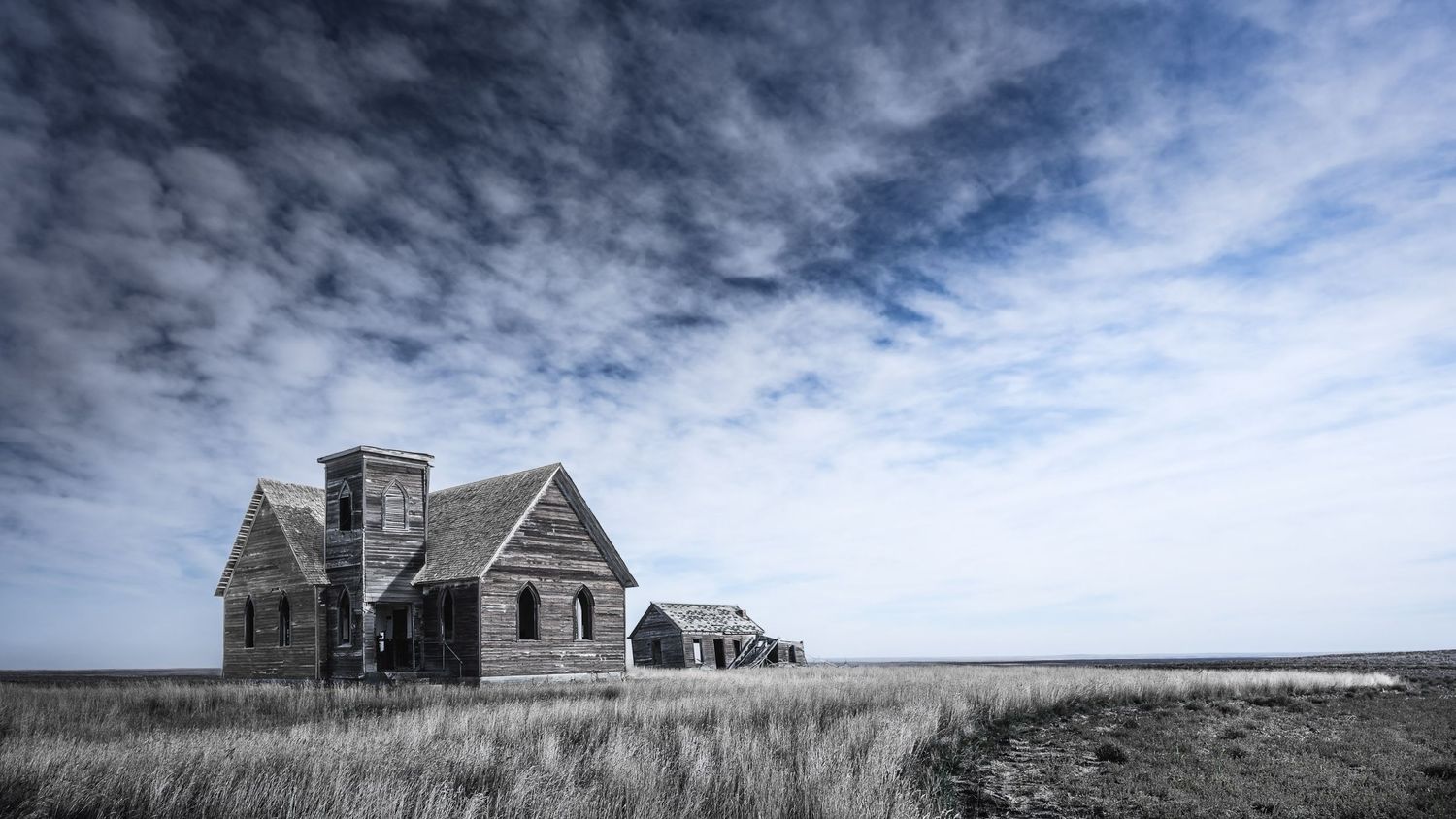 Abandoned wooden farmhouse deteriorates in vast prairie landscape beneath dramatic clouds.