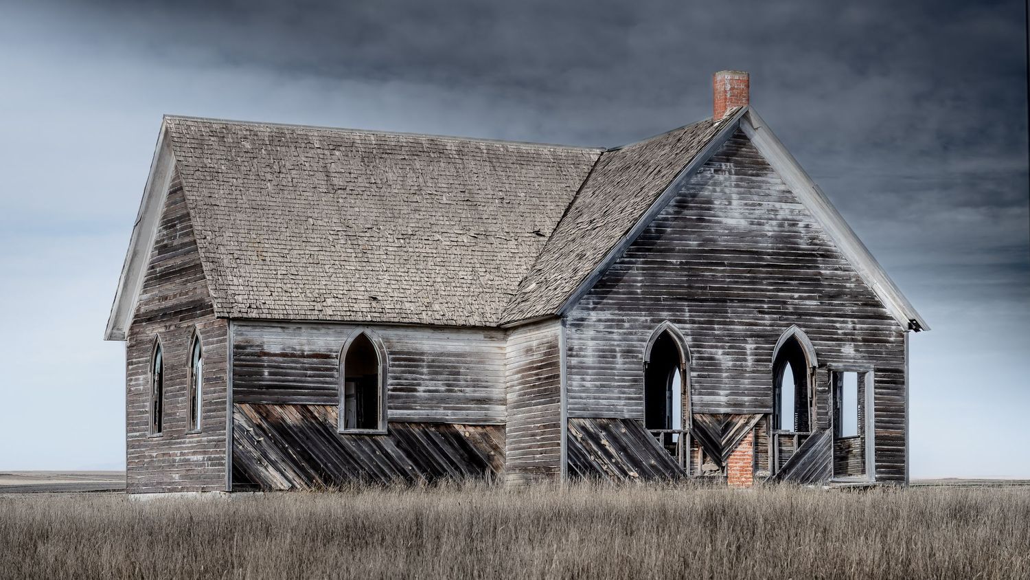 Dilapidated wooden church with Gothic windows sits abandoned in prairie grassland.