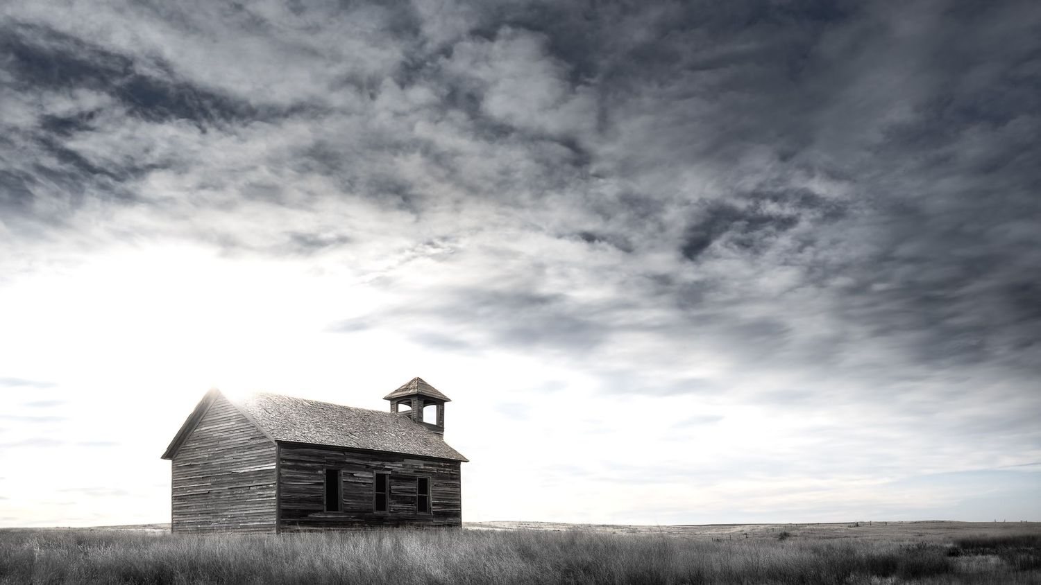 Weathered one-room schoolhouse with bell tower stands solitary against stormy prairie sky.