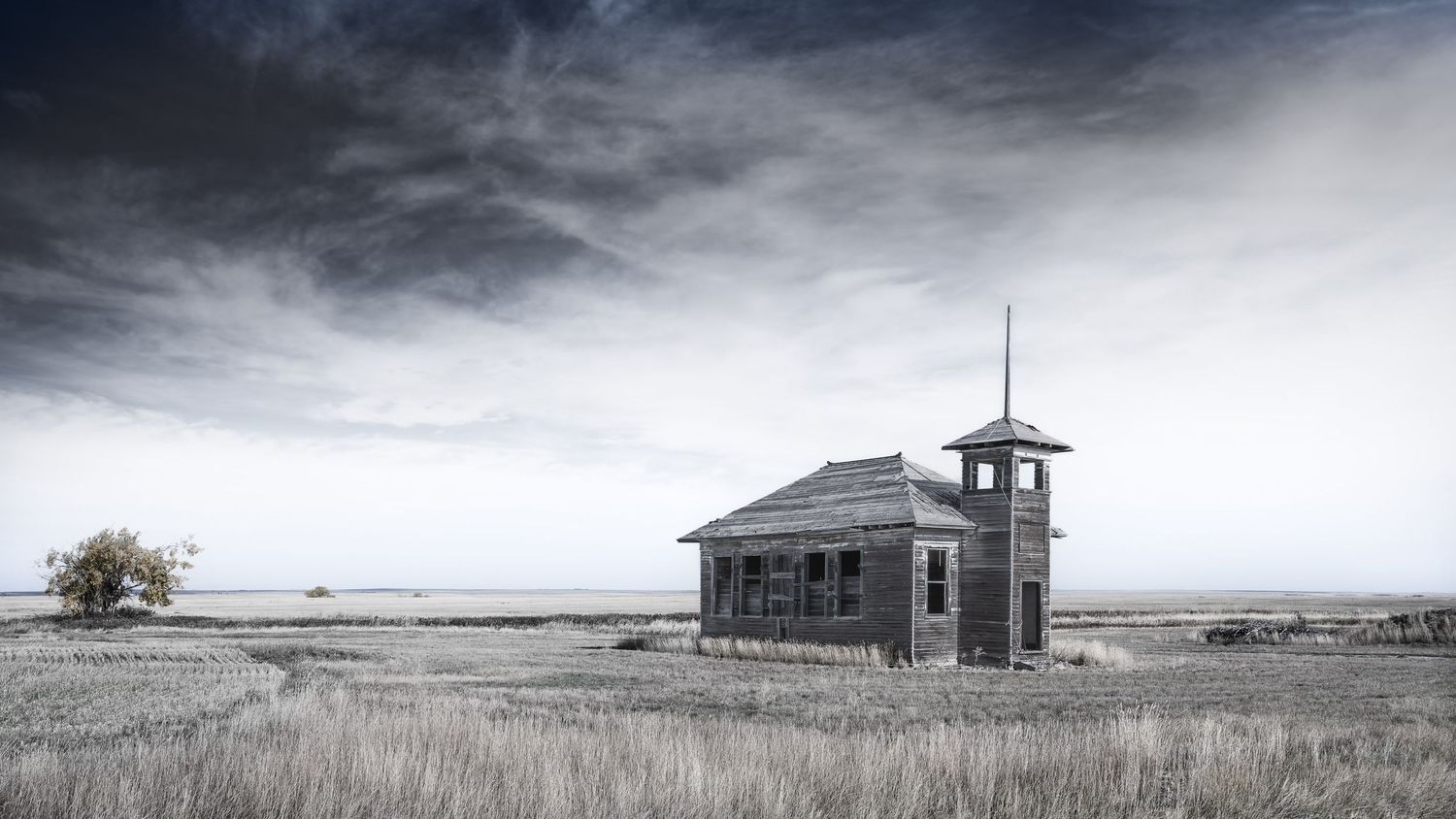 Abandoned one-room schoolhouse stands alone on prairie under dramatic cloudy sky.