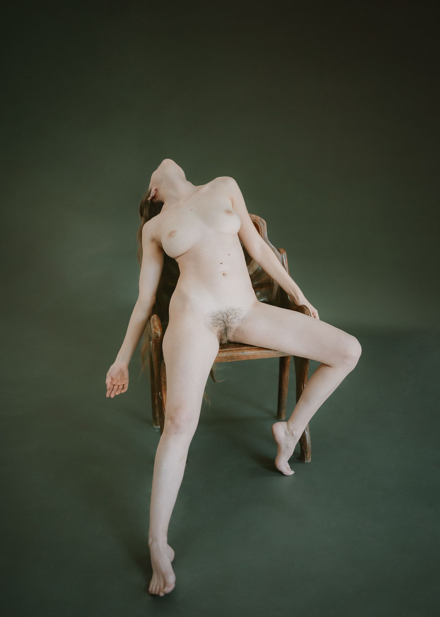 Artistic nude pose on wooden chair against muted green studio backdrop.