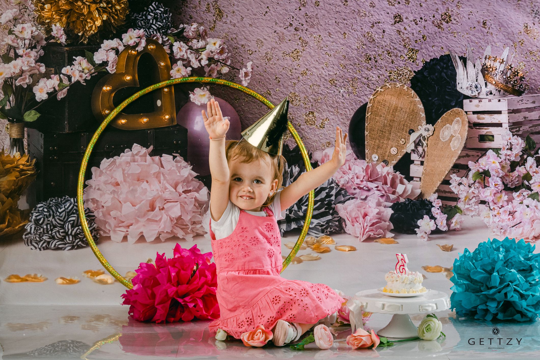 Colorful first birthday cake smash photo session with pink and teal decorations, gold hoop, and festive paper flowers.