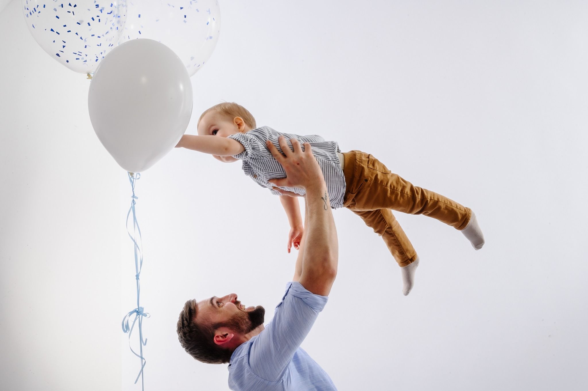 Joyful playtime scene with adult lifting child while white balloon floats nearby against light backdrop.