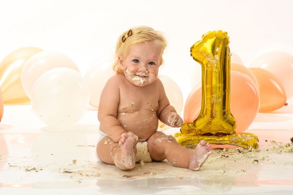A baby sits on a white floor with a gold number 1 balloon and pink and peach balloons in the background.