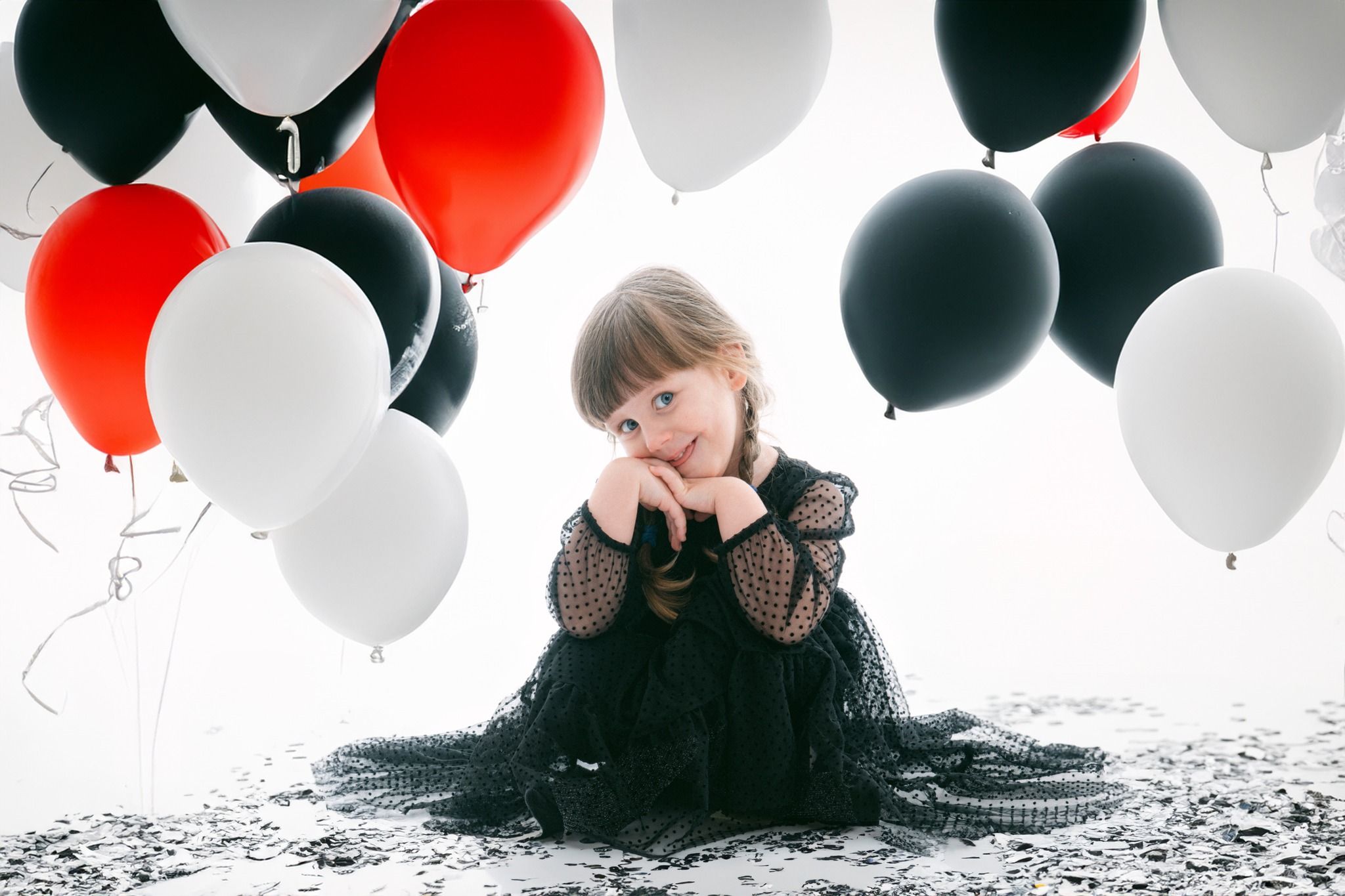 Child in black tulle dress poses with red, black and white balloons against white background.