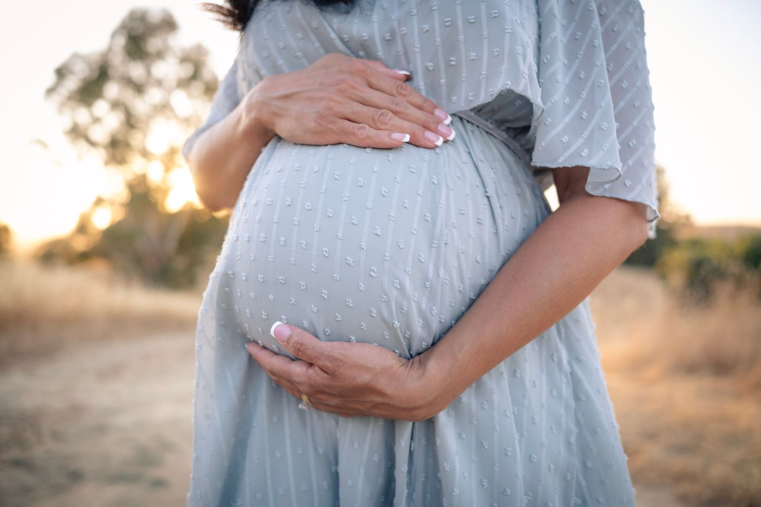 Hands gently cradle a pregnant belly in a light blue dress against a golden sunset backdrop outdoors.