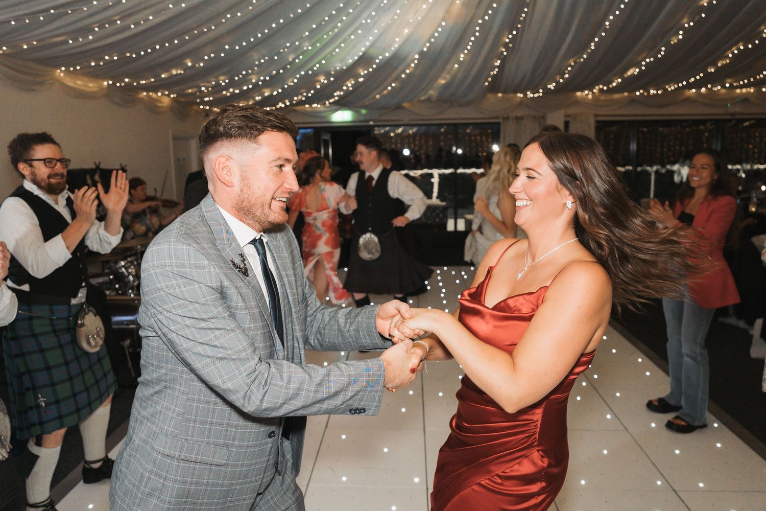 Two people dancing together at an indoor reception with string lights overhead and guests watching in the background.