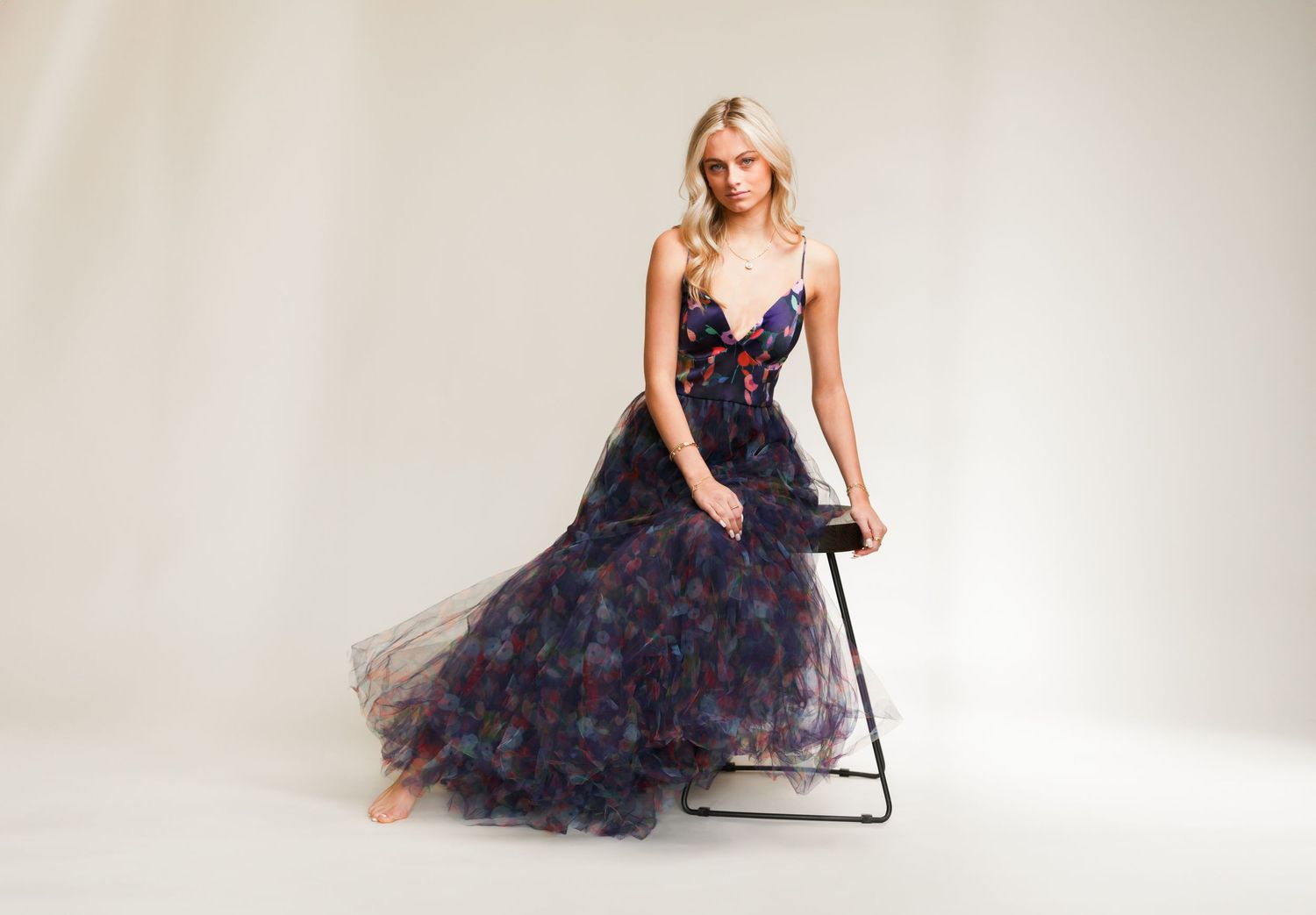 Elegant model in dark floral tulle gown with deep v-neckline poses elegantly on minimalist chair against beige backdrop.