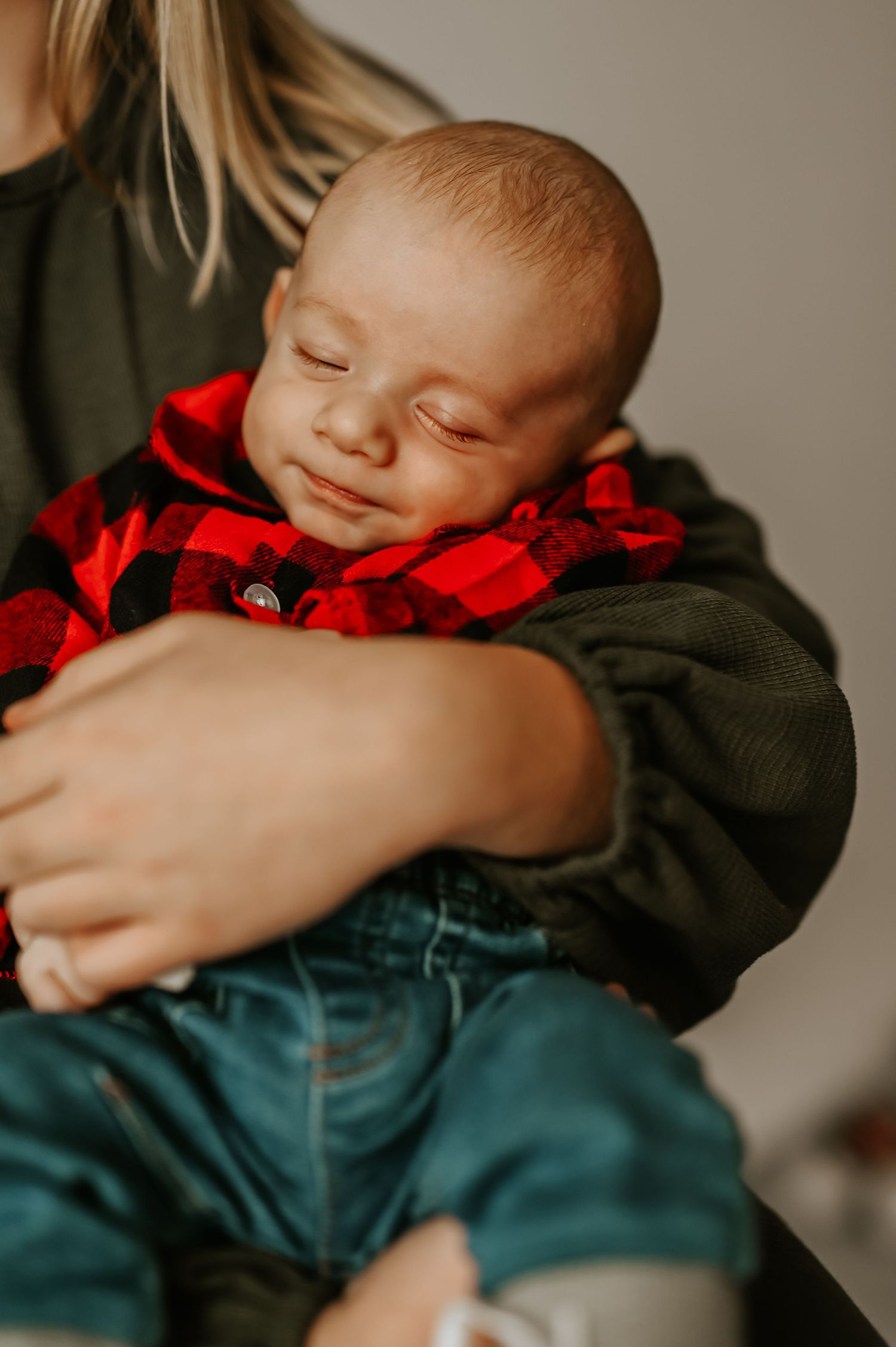 A newborn baby in red plaid shirt and jeans sleeping peacefully in big sisters arms during a Holiday Mini Session in Hartland, Wisconsin