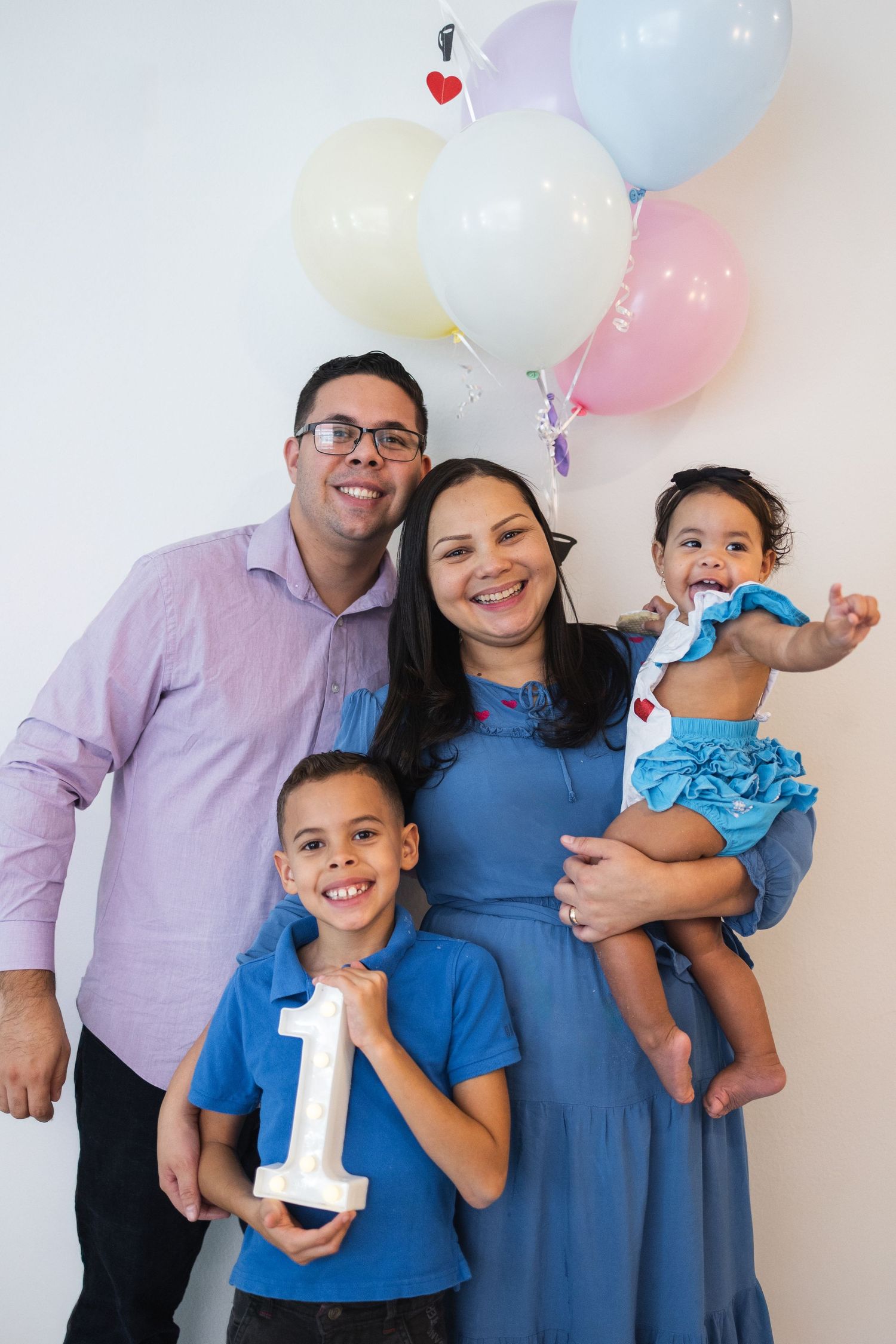 Family celebration with balloons as young children hold number one props against white wall.
