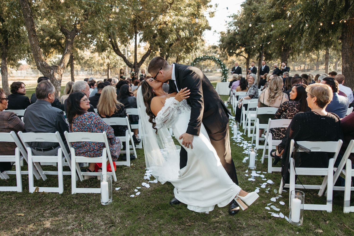 A romantic outdoor wedding ceremony in a garden setting with white chairs and guests watching the couple share a kiss.