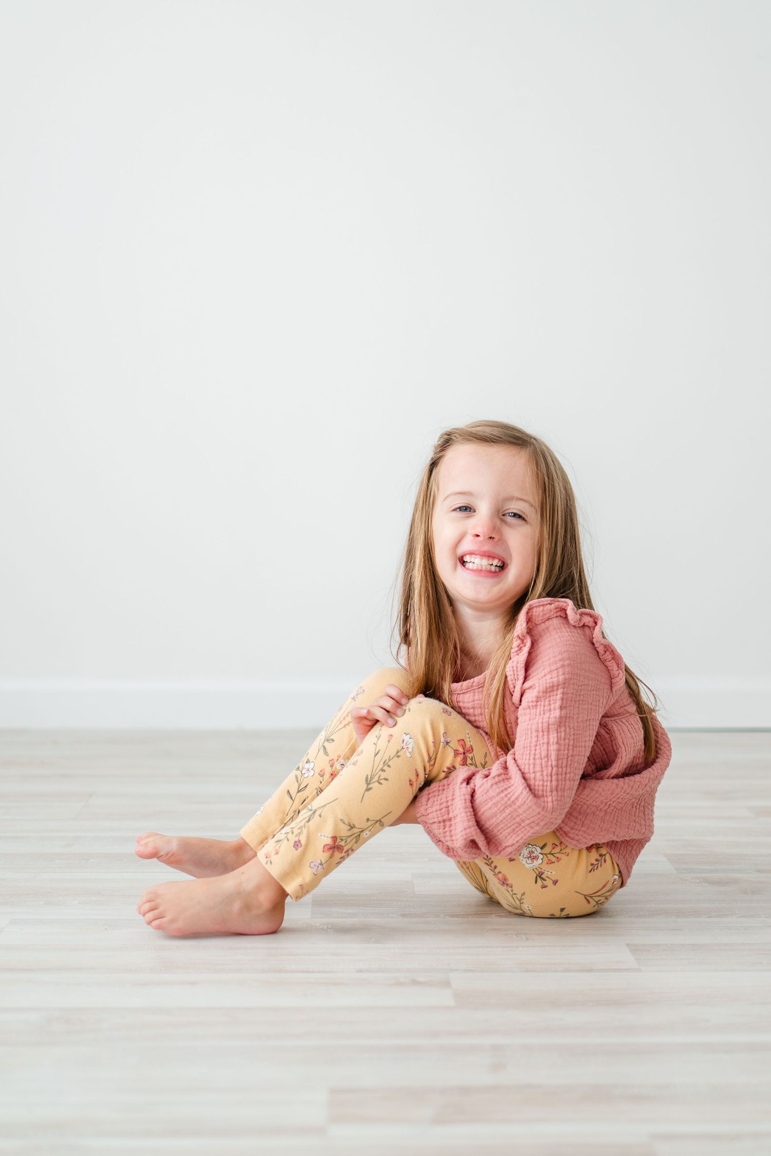 A young child wearing pink and yellow floral pajamas sits on a light wooden floor against a white wall.