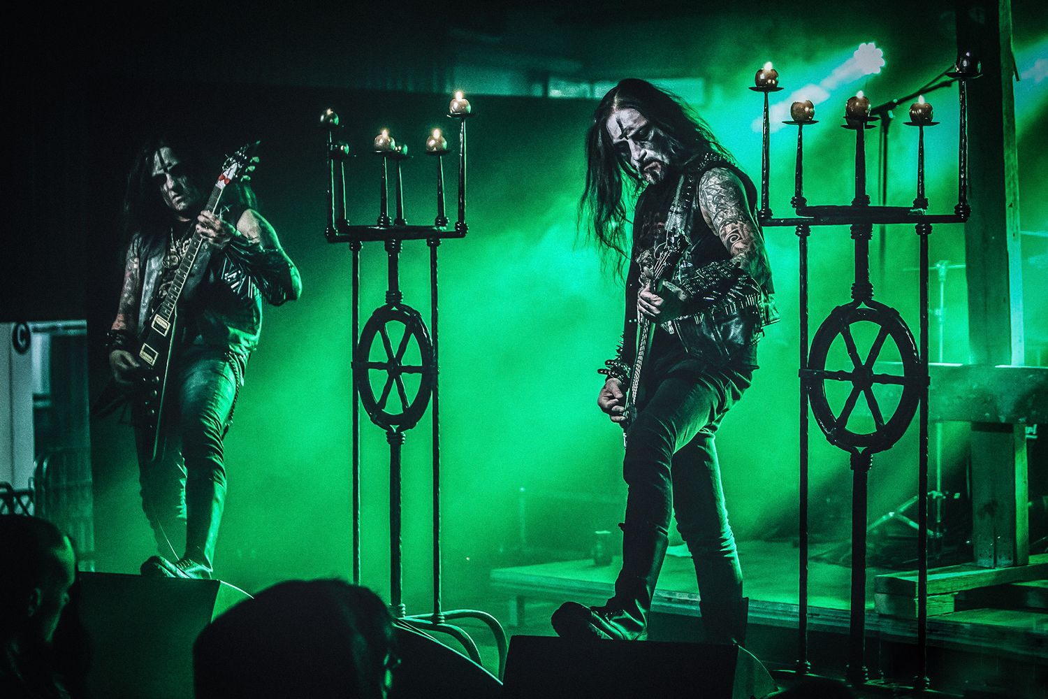 Metal band performs on stage with green lighting and dramatic medieval-style torch displays.