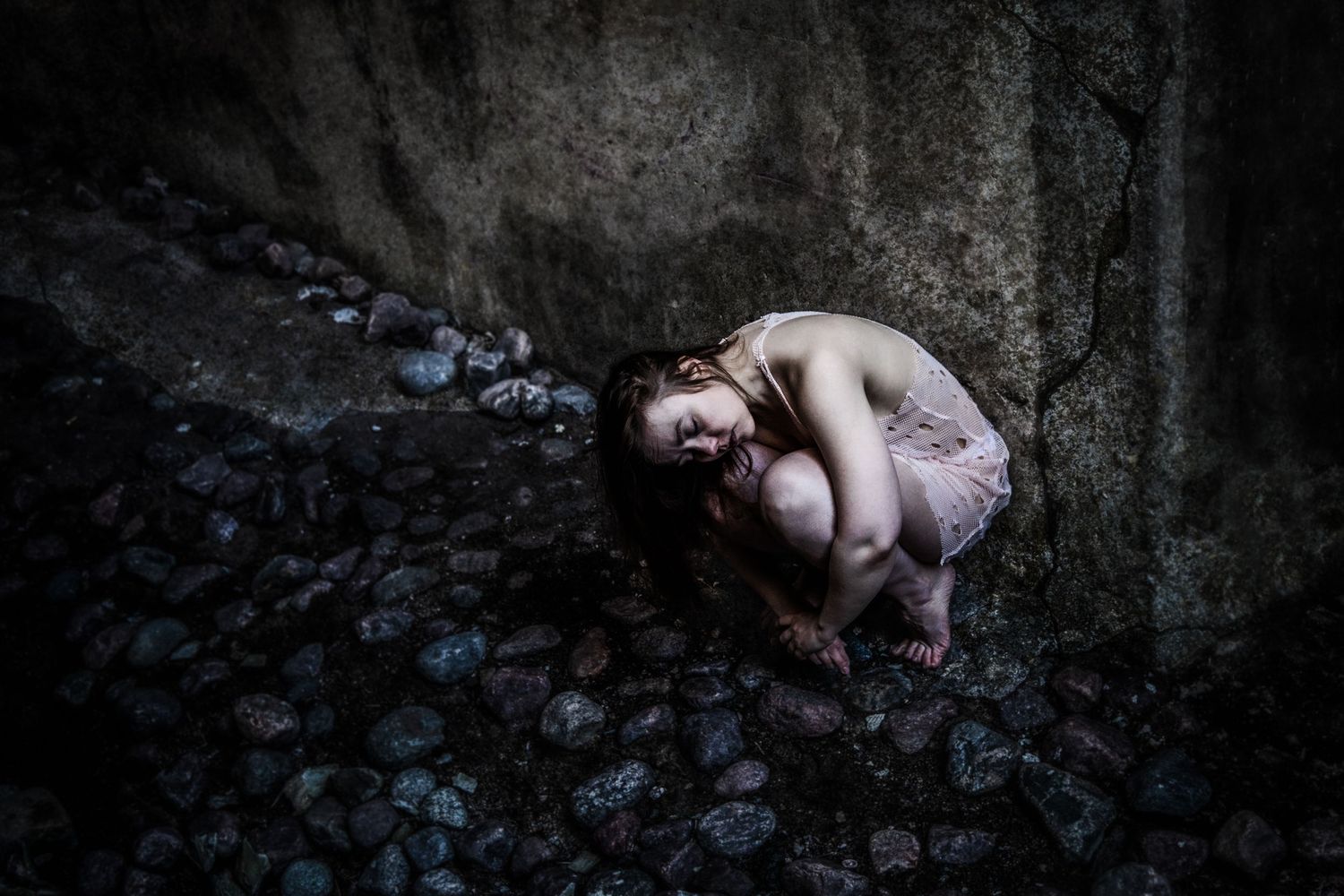 Artistic dark photograph showing a figure crouched among blue wildflowers against a stone wall in moody lighting.