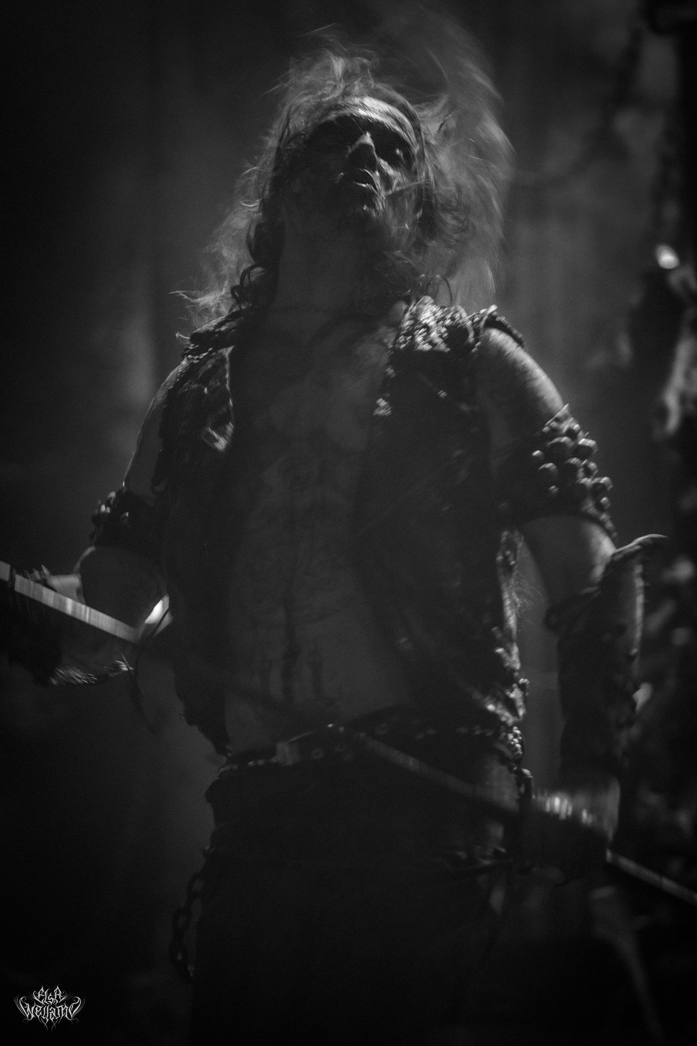 Black and white dramatic performance photo showing a musician backlit through smoke on a dark stage.