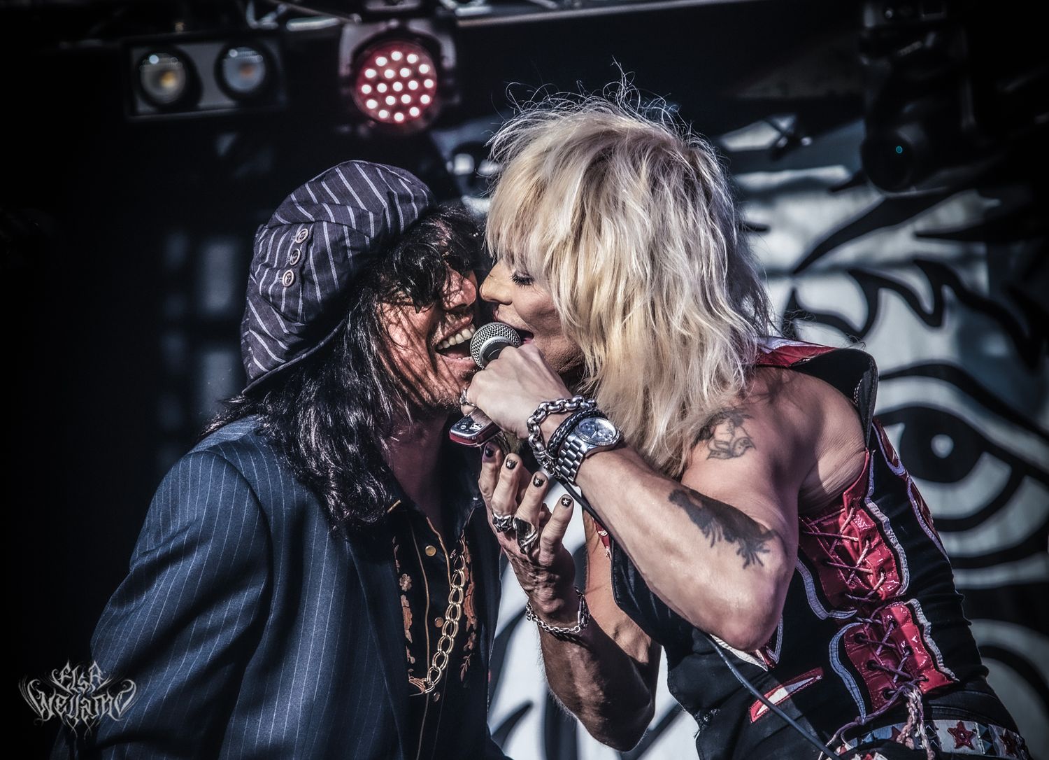 Two performers share an intense moment on stage during a rock concert with dramatic lighting and backdrop.