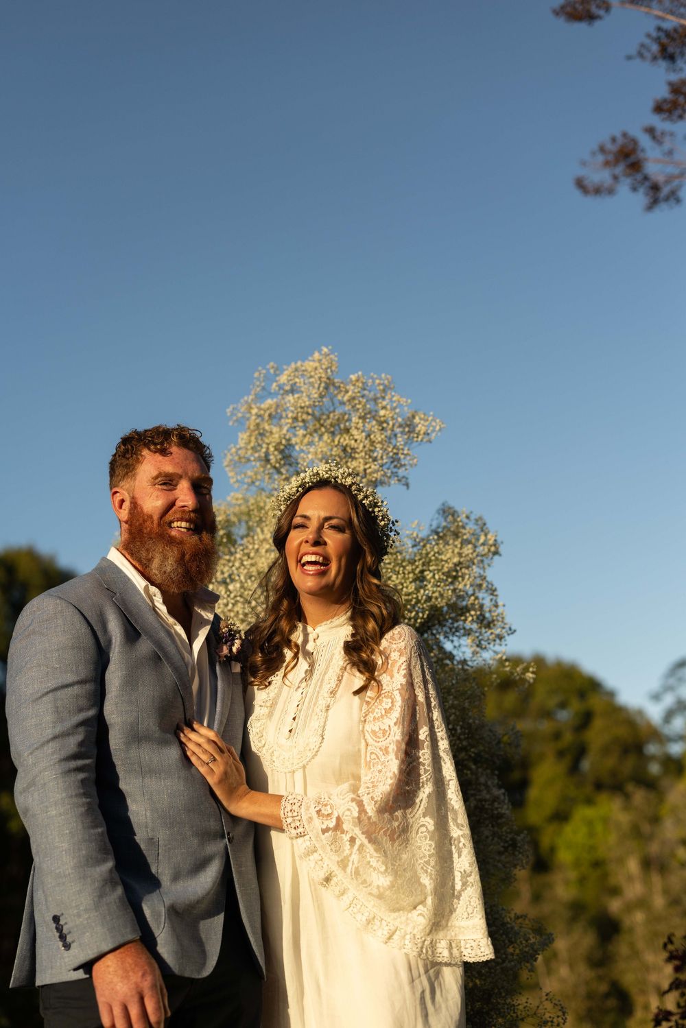 A couple laughs together while wearing formal attire outdoors with trees and blue sky in the background.