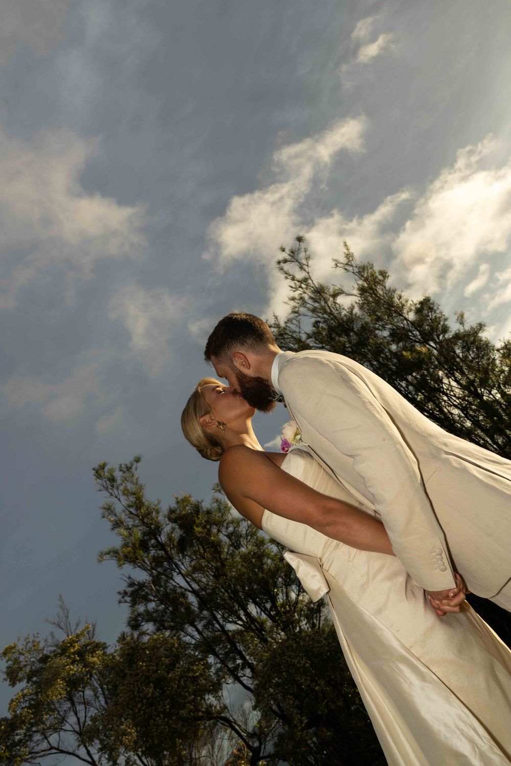 A romantic couple in light-colored attire share a tender kiss against a dramatic cloudy sky during a wedding ceremony.