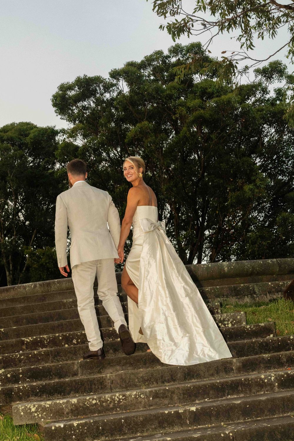 A couple in wedding attire sits together on concrete steps outdoors with trees in the background at sunset.