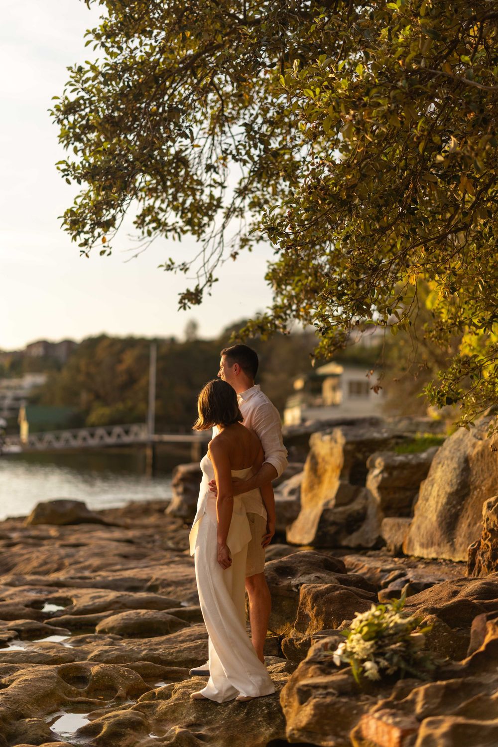 A couple embraces during golden hour on a rocky shoreline with trees and water in the background.
