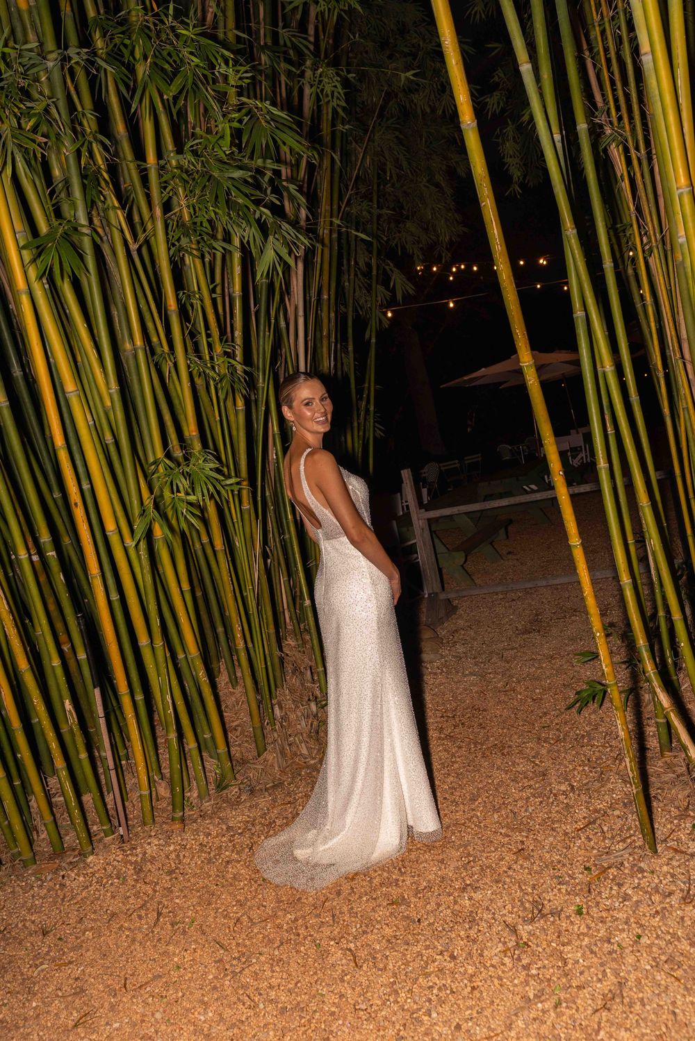 A person in a white dress poses among tall bamboo stalks at night in a garden setting.