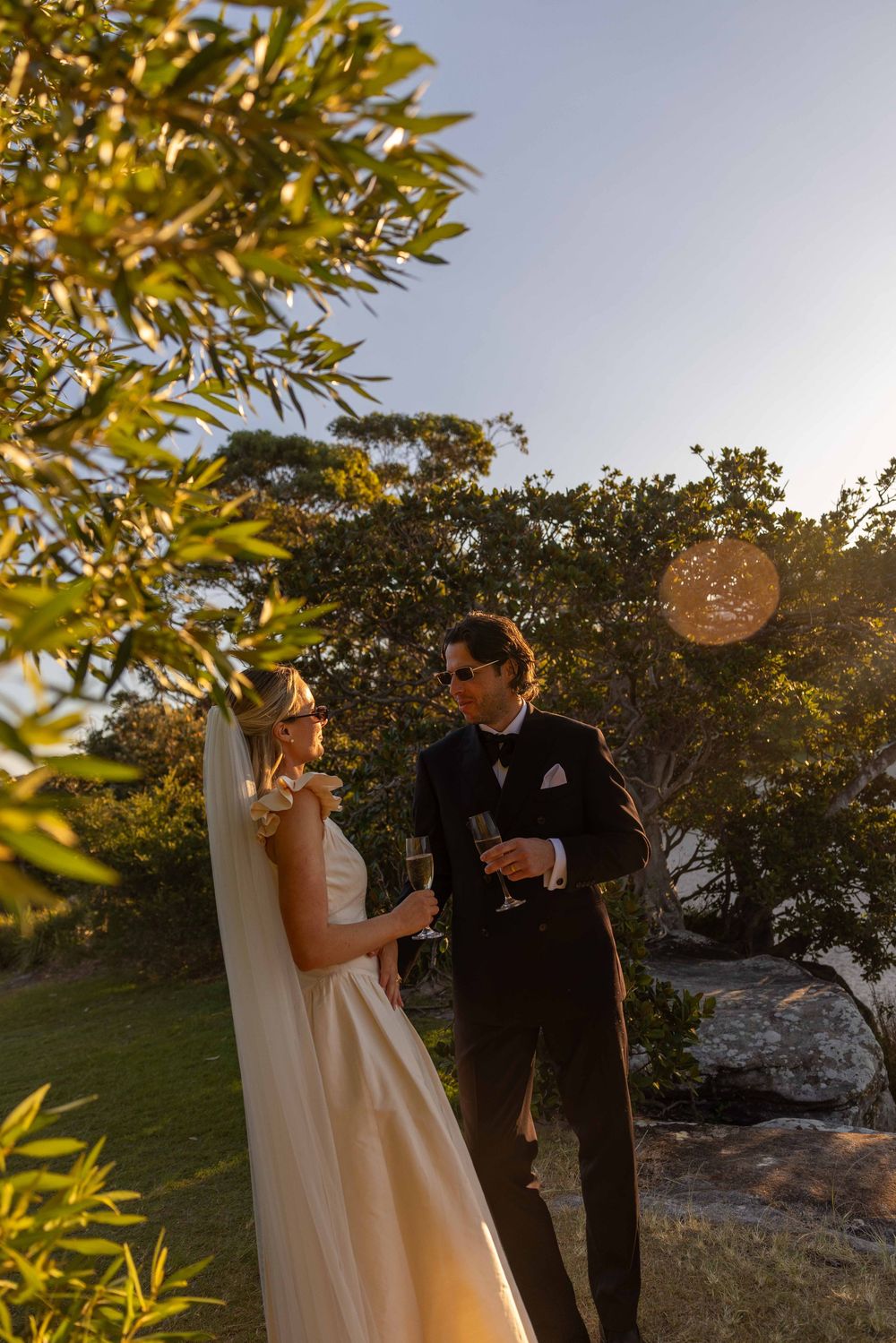 Bride and groom toast with champagne at golden hour outdoor wedding ceremony surrounded by lush greenery.