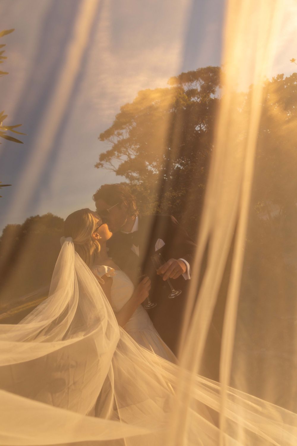 Golden hour wedding photo of bride and groom through flowing veil, sun rays illuminating romantic outdoor ceremony.