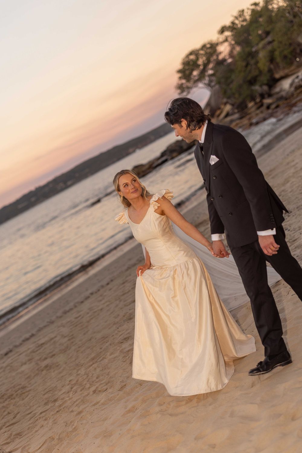 Bride and groom walking hand in hand on a sandy beach at sunset, bride in ivory gown, groom in black suit.