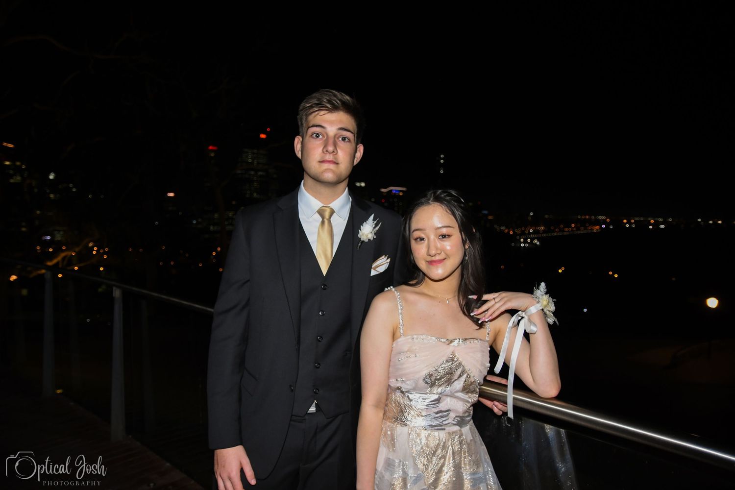 A formal couple poses at night on a balcony overlooking city lights in elegant evening attire.