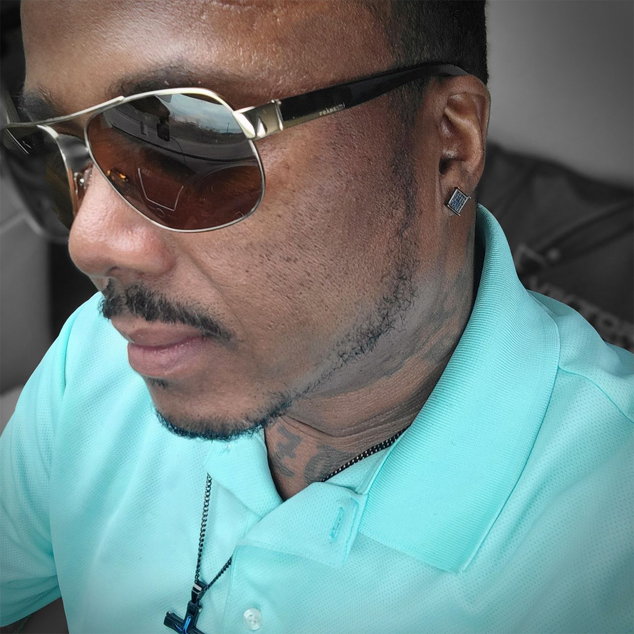 Profile view of someone wearing sunglasses and a turquoise polo shirt with neck tattoos visible in a close-up shot.