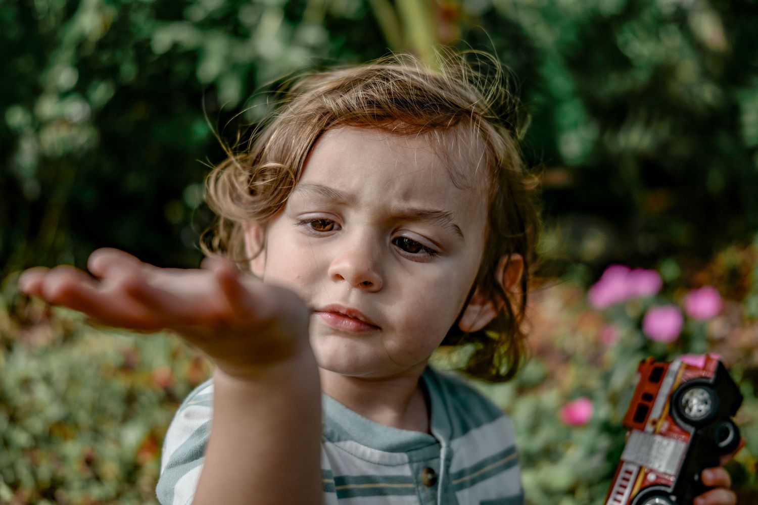 A small child reaches out their hand while standing in a lush garden with pink flowers in the background.
