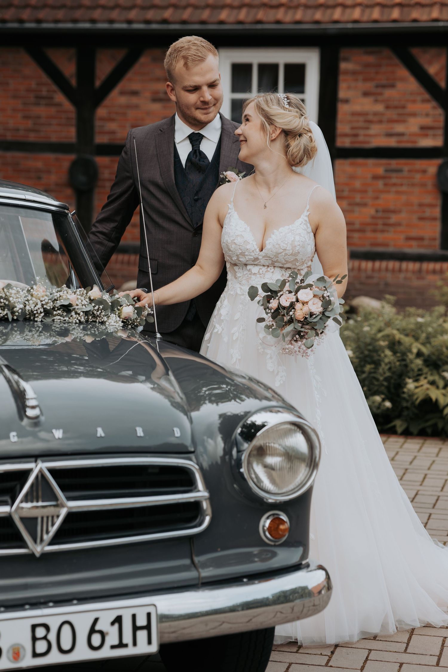 A couple in wedding attire stands beside a vintage car at a brick building, with the bride holding a pink bouquet.