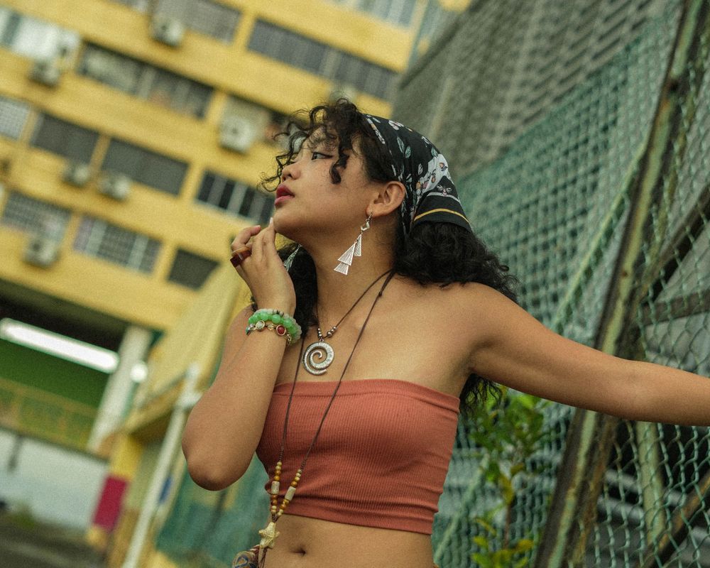 Urban portrait of woman in orange top with headphones against yellow building and fence.