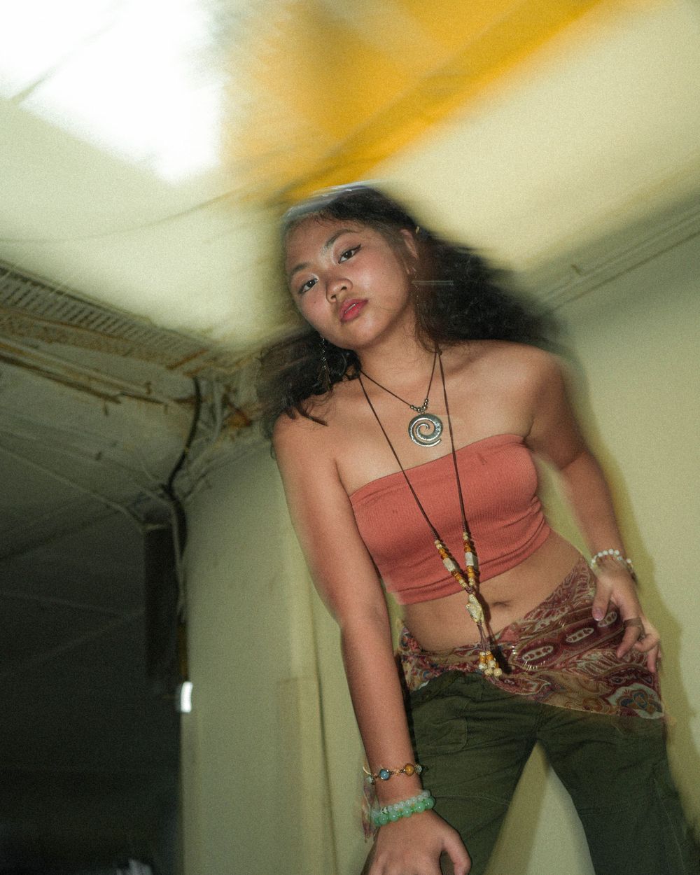 Artistic portrait of woman in orange top and jewelry against blurred interior background.