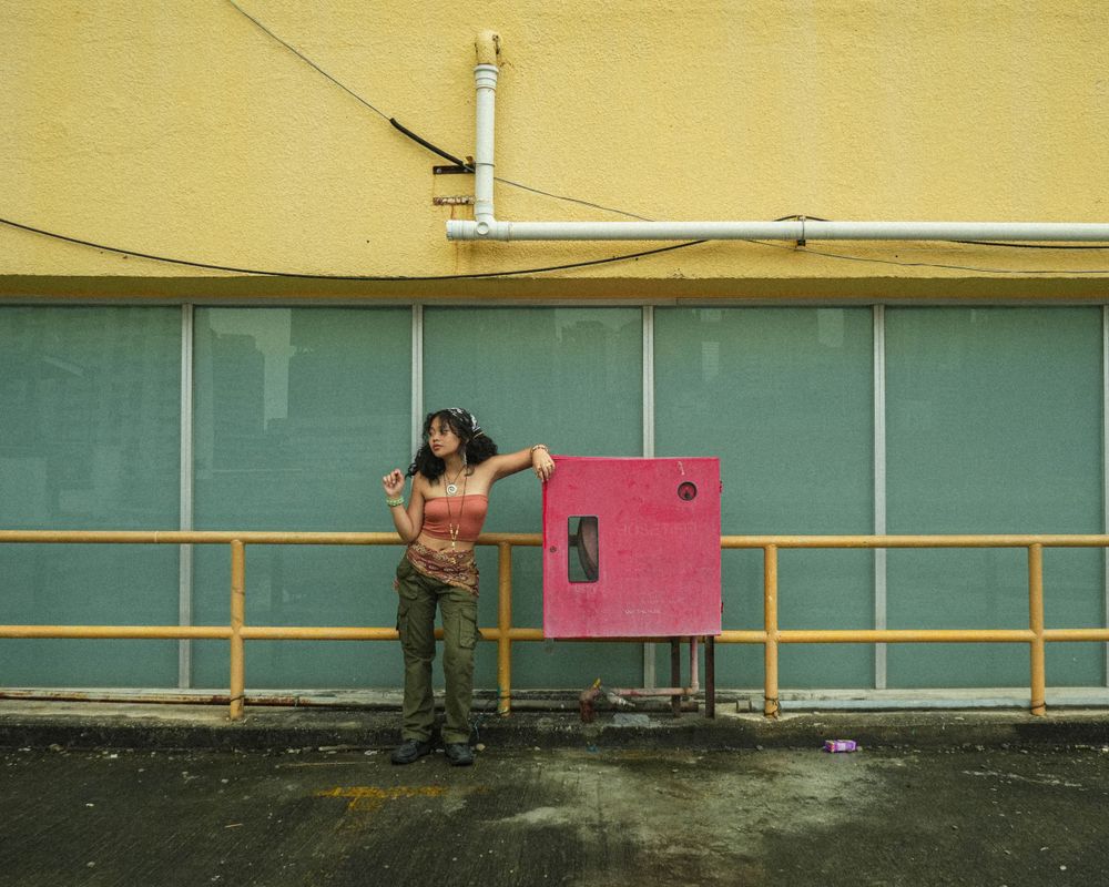 Woman in casual attire poses against turquoise wall with pink phone booth.