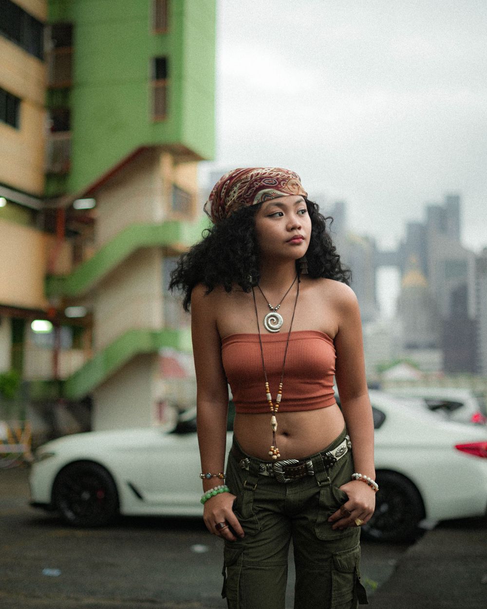 Street fashion photography with city skyline and luxury cars in background.