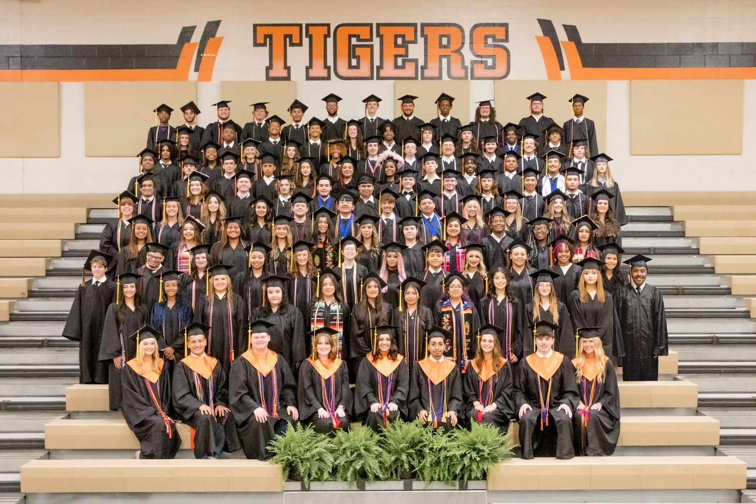 Large group of graduates wearing black caps and gowns posed on bleachers under a Tigers banner.
