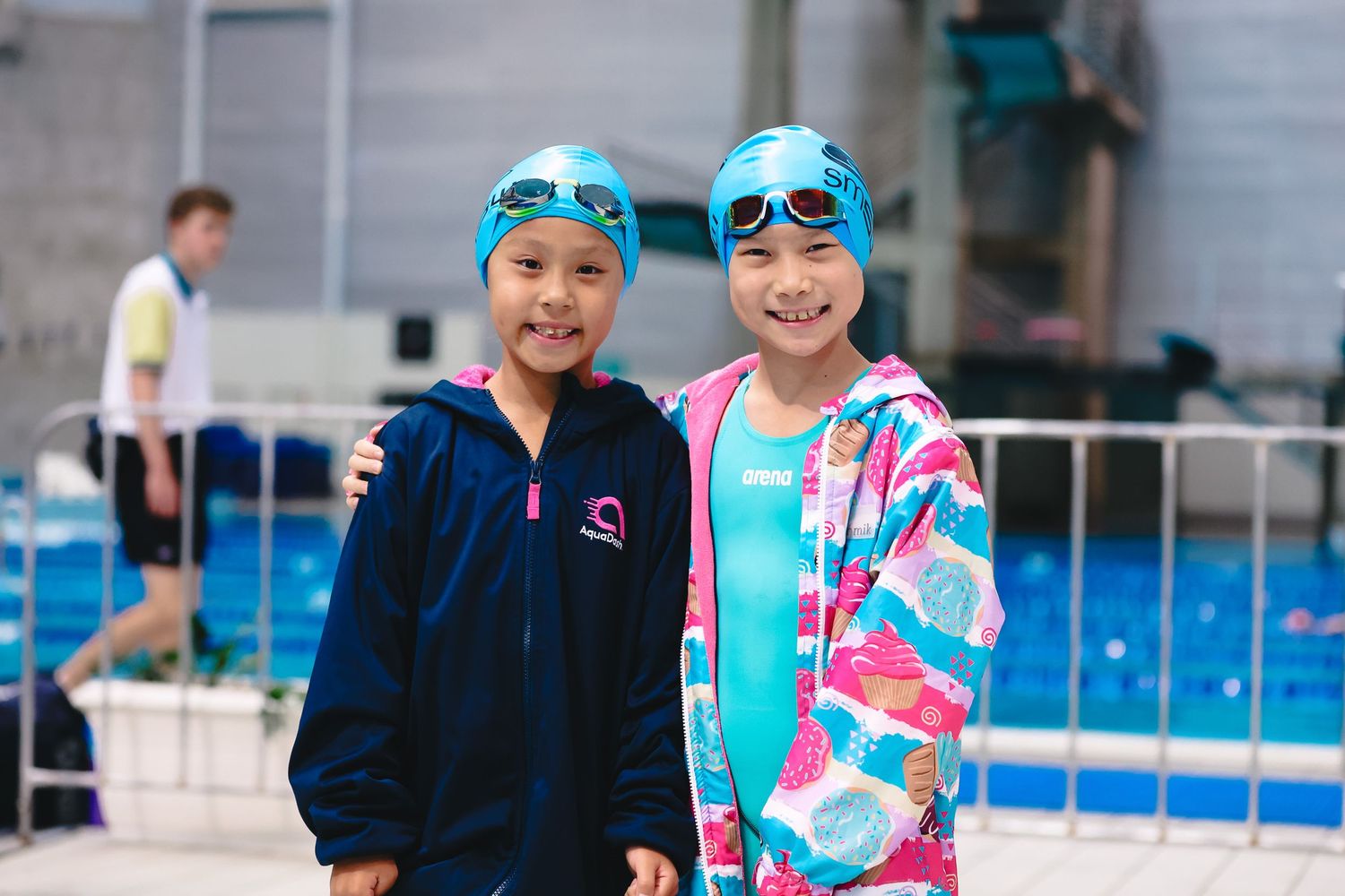 Two young swimmers wearing swim caps and colorful jackets stand together by a pool.