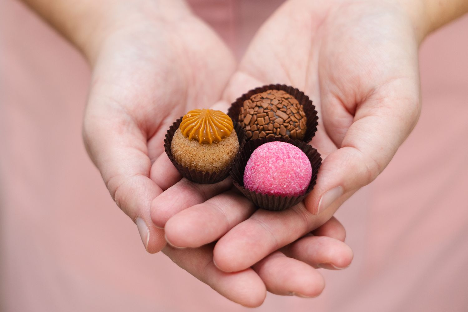 Three colorful chocolate truffles being held in open palms against a pink background.