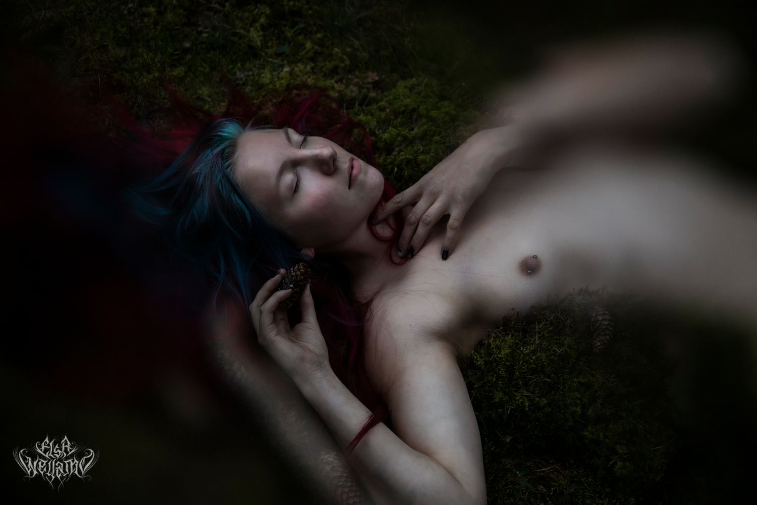 Dramatic artistic portrait shows resting figure with blue hair lying on grass in soft natural lighting.