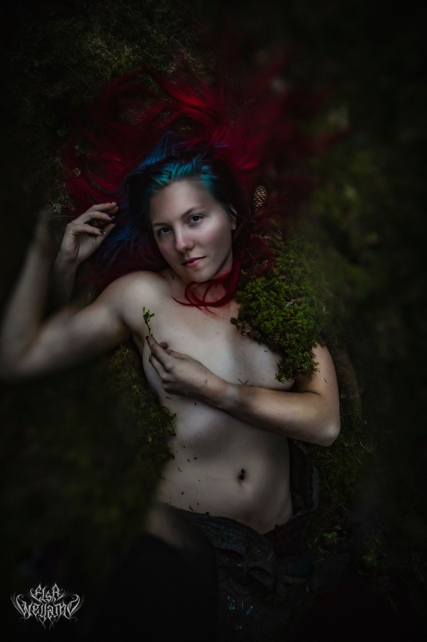 Artistic dark forest photoshoot with vibrant red and blue hair details against moody green background.